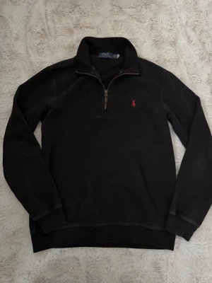 Svart half zip tröja Polo Ralph Lauren - Klassisk svart långärmad tröja från Polo Ralph Lauren med half zip och röd broderad logga på bröstet. Tröjan har hög krage och är gjord i mjuk bomull, perfekt för lager på lager under höst och vinter.