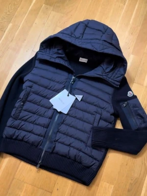 Mörkblå pufferjacka från Moncler - Säljer en mörkblå pufferjacka från Moncler med huva och ribbade muddar. Jackan har quiltad front, dragkedja och fickor med knappstängning. Ärmarna är stickade och har en liten ficka med dragkedja på ena armen. Perfekt för kyliga dagar.