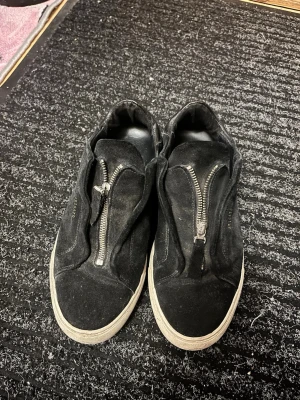 Svarta sneakers från Axel Arigato, strl 41 - Svarta sneakers från Axel Arigato i mocka med cool dragkedja framtill istället för klassisk snörning. Snygg vit sula och diskret guldfärgad logga på sidan. Skorna har en stilren och modern look som passar till det mesta.