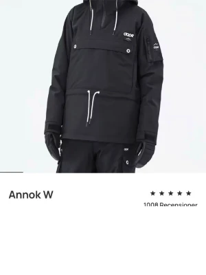 Svart jacka från Dope Annok - Säljer en svart vindjacka från Dope Annok med huva och vita snören. Jackan har en stor ficka framtill, dragkedja vid halsen och logga på bröstet och ärmen. Perfekt för kalla dagar och outdoor-äventyr.