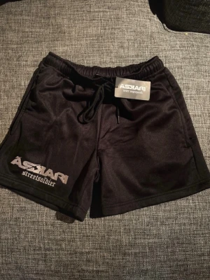 Svarta shorts från Askari Streetsoldier - Sprillans.nya Svarta shorts från Askari Streetsoldier med tryck på ena benet och siffran 83 på andra. De har elastisk midja med snörning och är gjorda i ett mjukt, syntetiskt material som passar perfekt till träning eller chill. Snygg och sportig look.