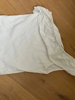 Vit oneshoulder topp från Kappahl - Säljer en vit oneshoulder topp från Kappahl i ribbad bomull med bred volangkant i broderad spets. Toppen har en axel och fina detaljer som ger en romantisk vibe. Perfekt till sommaren och lätt att matcha med jeans eller kjol.