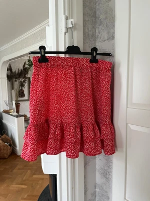 Röd blommig volangkjol från ONLY - Supersöt röd kortkjol från ONLY med små vita blommor och volangkant nertill. Kjolen har resår i midjan för en bekväm passform och är perfekt för varma dagar. Lätt och luftig, passar till sneakers eller sandaler.