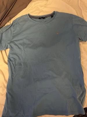 Ljusblå t-shirt från Gant - Ljusblå t-shirt från Gant med klassisk rund hals och liten broderad logga på bröstet. Tillverkad i mjuk bomull som känns skön mot huden. Enkel och stilren design som passar till det mesta.