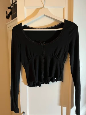 Svart croppad långärmad topp med spets - Säljer en svart croppad topp med långa ärmar. Toppen har spetsdetaljer runt ringningen och framtill, samt en liten rosett vid urringningen. Volangkant nedtill ger en söt touch. Perfekt till jeans eller kjol för en trendig look.