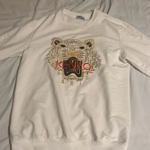 Vit sweatshirt med tiger från Kenzo - Säljer en vit sweatshirt från Kenzo med ikoniskt broderat tigermotiv och Kenzo Paris-text i rött och guld på bröstet. Tröjan har rund hals, långa ärmar och ribbade muddar. Perfekt statement piece för dig som gillar streetwear och snygga detaljer.