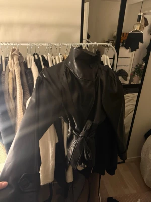 skinn trenchcoat med bälte - Säljer en svart trenchcoat i skinnimitation med hög krage och knytbälte i midjan. Jackan har långa ärmar och en clean, stilren look som passar perfekt till lager på lager. Snygg detalj med knappar vid ärmslut och axlar.