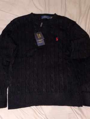  Polo Ralph Lauren tröja - Kabelstickad mörkblå tröja från Polo Ralph Lauren med klassisk röd logga broderad på bröstet. Tröjan har rund halsringning, ribbade muddar och är tillverkad i mjuk ullblandning. Perfekt för dig som vill ha en stilren och tidlös look.