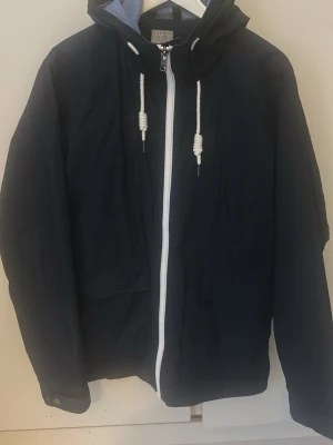 Mörkblå vårjacka från Etirel - Säljer en mörkblå vindjacka från Etirel i storlek M. Jackan har vit dragkedja, huva med vita snören och två stora fickor framtill. Materialet är lätt och syntetiskt, perfekt för blåsiga dagar. Enkel och stilren design som funkar till det mesta.