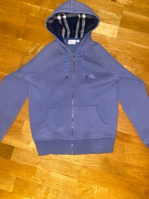 Burberry blå zip-hoodie - Original Burberry hoodie i blå färg med dragkedja framtill. Tröjan har Burberrys klassiska rutiga foder i huvan och en diskret broderad logotyp på bröstet. Tillverkad i mjukt och bekvämt material som passar perfekt för vardag, fritid eller som ett stilrent lager under svalare dagar. En tidlös och exklusiv huvtröja från ett välkänt premiumvarumärke.