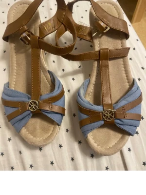 Blå och bruna kilklackssandaler strl 39 - Säljer ett par sandaler med kilklack i flätad look. Ovandelen är i ljusblå textil och bruna remmar i syntet, dekorerade med en metallknapp framtill. Remmarna går runt ankeln och har spänne. Perfekta för sommaren och ger en chill vibe till din outfit.