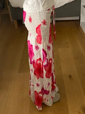 Vit långkjol med stora blommor - Säljer en flowy långkjol i vitt med stora blommiga mönster i rött och rosa. Kjolen har smock i midjan och knytband för extra passform. Materialet känns lätt och luftigt, perfekt för varma dagar. Blommönstret ger en färgstark och somrig vibe. Fungerar även som en klänning. Har ett litet hål längst ner, men syns inte när den e på och går enkelt att sy ihop 🩷🩷🩷