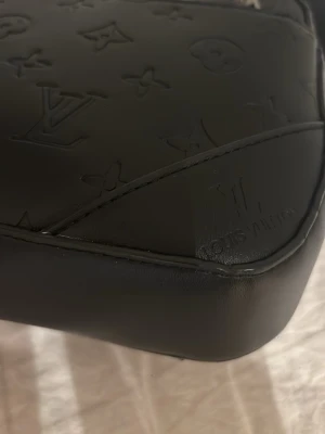 Svart axelväska  - Säljer en svart axelväska i skinn med präglat monogrammönster. Väskan har bred justerbar axelrem, dragkedja med metall-detaljer och stilren design. Perfekt för dig som vill ha en lyxig och praktisk väska med ikonisk look.