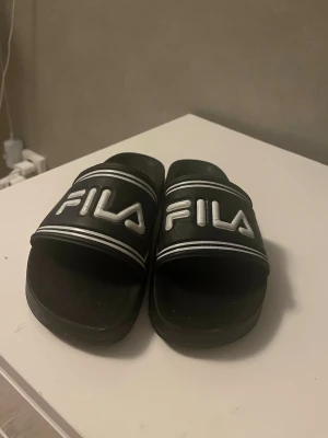 Svarta Fila sandaler med vit logga - Säljer ett par svarta Fila sandaler med bred rem och stor vit Fila-logga framtill. Skorna har en platt sula och vita ränder som detalj på remmen. Perfekta för sommaren eller att glida runt hemma i.
