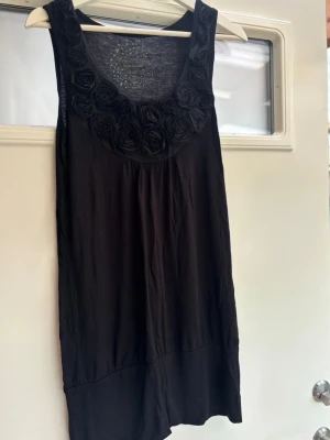 Svart ärmlös topp med rosdetaljer - Säljer en svart ärmlös topp från KappAhl med snygga tygrosor runt ringningen. Toppen har en lös passform och är gjord i mjukt bomullsmaterial. Perfekt att styla med jeans eller kjol för en cool look.