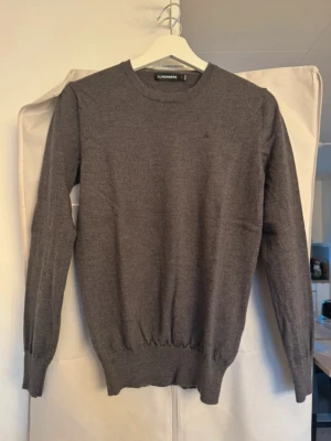 Grå stickad tröja från J.Lindeberg - Mjuk och stilren grå stickad tröja från J.Lindeberg. Klassisk rund halsringning och ribbade muddar vid ärmslut och nederkant. Perfekt för dig som gillar en enkel men snygg look.