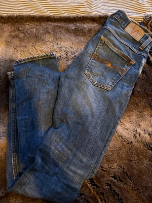 Blå Nudie Jeans Slim Straight - Säljer ett par klassiska blå jeans från Nudie Jeans med snygga slitningar och kontrastsömmar. Modellen har en rak och smal passform, fem fickor och detaljerad bakficka med nudie loggan.
