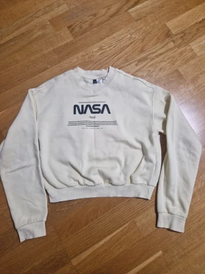 Vit NASA sweatshirt från H&M XS - Säljer en vit sweatshirt från H&M i storlek XS med stort svart NASA-tryck på bröstet. Tröjan har rund hals, långa ärmar och ribbade muddar vid ärmslut och nederkant. Perfekt för dig som gillar rymd och streetstyle.
