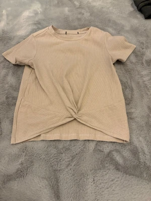 Beige ribbad topp med knut framtill - Säljer en beige ribbad topp med korta ärmar och rund hals. Toppen har en snygg knutdetalj framtill som ger en chill vibe. Materialet är mjukt och stretchigt, perfekt för en avslappnad look.