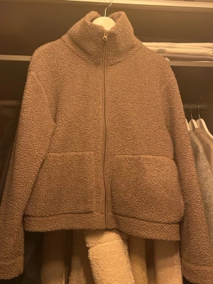 Beige teddyjacka med dragkedja - Mysig beige teddyjacka med hög krage och hel dragkedja framtill. Jackan har två stora fickor och är tillverkad i fluffigt teddy-material som håller dig varm under kyliga dagar. Perfekt för lager på lager och chill vardagsstil.