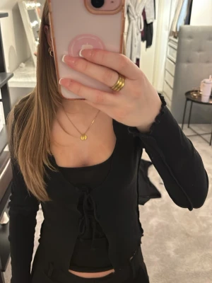 Svart kofta med snörning - Säljer en svart kofta med långa ärmar och snygg snörning framtill. Koftan har vågiga kanter vid ärmslut och nederkant som ger en trendig look. Perfekt att slänga över ett linne eller topp för en chill vibe.