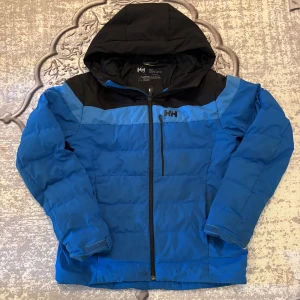 Blå pufferjacka från Helly Hansen - Säljer en blå och svart pufferjacka från Helly Hansen med huva och dragkedja framtill. Jackan har en bröstficka med dragkedja, justerbara ärmslut med kardborre och meshficka på insidan. Perfekt för kalla dagar och outdoor vibes. Inte sliten eller något men bara att färgen har ändrats. Riktig varmt och skönt. Köpte runt 3000.  Priset är inte i hugget så kan diskutera 🙂