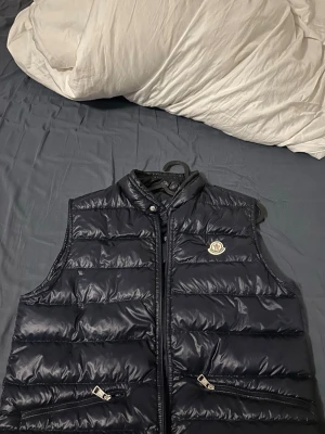 Marinblå  väst från Moncler - Säljer en svart dunväst från Moncler med klassisk quiltad design och hög krage. Västen har dragkedja framtill, två fickor med dragkedja och Moncler-logga på bröstet. Perfekt för lager-på-lager och snygg till både hoodie och t-shirt. Köpt för 6000 på merchsweden i 2025 somras och såljer för 4600. Storlek 3