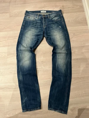 Scotch & Soda Ralston blå jeans - Säljer ett par blå Scotch & Soda Ralston jeans med snyggt slitna detaljer och klassisk femficksmodell. Jeansen har en  slim passform och ett brunt läderpatch bak i midjan. Materialet är robust denim i bomull och färgen är tvättad blå med ljusare partier.