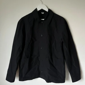 Svart overshirt från ARKET - Svart skjortjacka från ARKET i stilren design med klassisk krage och knäppning framtill. Jackan har två stora fickor och är tillverkad i bomull. Perfekt för lager-på-lager och passar dig som gillar minimalistisk stil. 4A10