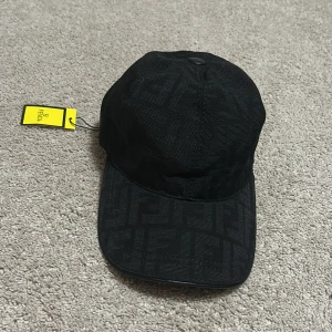 Svart Fendi keps med logomönster - Svart keps från Fendi med ikoniskt ton-i-ton logomönster över hela kepsen. Tillverkad i 100% bomull med justerbar rem bak. Snygg och stilren design som lyfter din outfit med exklusiv Fendi-vibe.