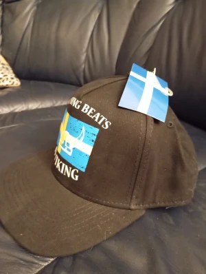 Svart keps med svensk viking-tryck - Cool svart keps med tryck av svensk flagga och vikingahjälm på framsidan. Texten 'Nothing beats a Viking' ger den extra attityd. Perfekt för dig som vill visa din svenska stolthet och vikinga-vibe. Justerbar baktill för skön passform.
