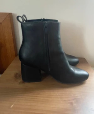 Svarta boots med klack och dragkedja - Snygga svarta boots med rund tå och stabil blockklack. Skorna har dragkedja på sidan och två öglor upptill för enkel påtagning. Tillverkade i skinnimitation med slät finish, perfekta för dig som gillar stilrena och klassiska skor.