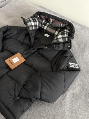 Burberry jacka  - Svart pufferjacka från Burberry med avtagbara ärmar, så du kan bära den som väst. Jackan har huva med klassiskt rutigt foder i svart, vitt och rött. Stora dragkedjefickor och Burberry-logga på ärmen. Perfekt för kalla dagar med stil.