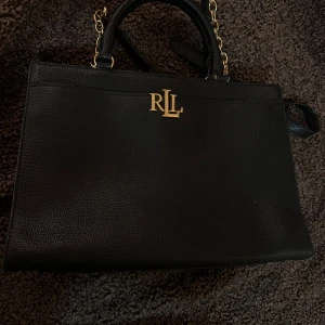 Svart handväska från Lauren Ralph Lauren - Snygg svart handväska i skinn från Lauren Ralph Lauren ÄKTA  med guldfärgade detaljer och kedja. Stilren och fyrkantig form med RLL-logga framtill. Väskan har praktiska handtag och dragkedja upptill för säker förvaring av dina grejer.