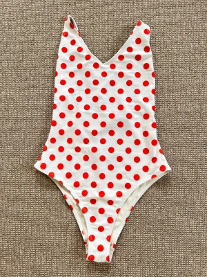 Vit baddräkt med röda prickar - Helt ny - Becksöndergaard - Supersnygg vit baddräkt från Becksöndergaard med röda prickar och smala axelband som korsas i ryggen. V-ringad framtill och öppen rygg med snörning för extra cool detalj. Perfekt för stranden eller poolen i sommar. Helt ny med taggar kvar. Tillverkad i 100% återvunnen polyester, vilket gör den både miljövänlig och skön att bära. 