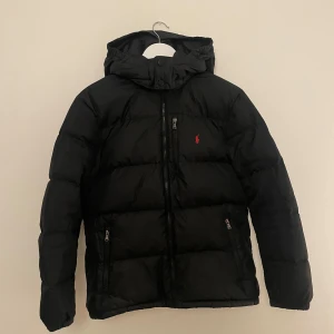 Svart pufferjacka från Polo Ralph Lauren - Svart pufferjacka från Polo Ralph Lauren med huva och dragkedja framtill. Jackan har två sidofickor med dragkedja och en liten röd Polo-logga på bröstet. Perfekt för kalla dagar och har en clean look..
