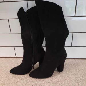 Svarta höga mockastövlar med klack - Snygga svarta stövlar i mocka med hög klack. Stövlarna har dragkedja på sidan för enkel på- och avtagning. Stilren design som går upp till knäet och ger en cool siluett. Perfekt för dig som vill ha en trendig look med extra höjd.