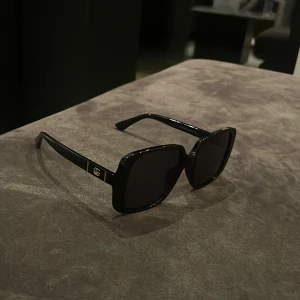 Svarta Gucci solglasögon, fyrkantiga - Snygga svarta solglasögon från Gucci med fyrkantiga, breda bågar och mörka linser. Guldiga detaljer med Gucci-logga på sidorna ger en lyxig vibe. Modellen är GG0638SK och bågarna är tillverkade i blank syntet. Perfekt för dig som vill sticka ut med stil.