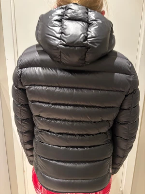 Svart Colmar dunjacka 2286N XS - Säljer en svart dunjacka från Colmar med huva och dragkedja framtill. Jackan är quiltad och har två fickor med dragkedja. Klassisk pufferstil med Colmar-logga på ärmen samt på ryggen. Modellen heter 2286N, stl 42 (motsvarar ca XS). Perfekt för kalla dagar. Mycket fint skick och 100% äkta. 