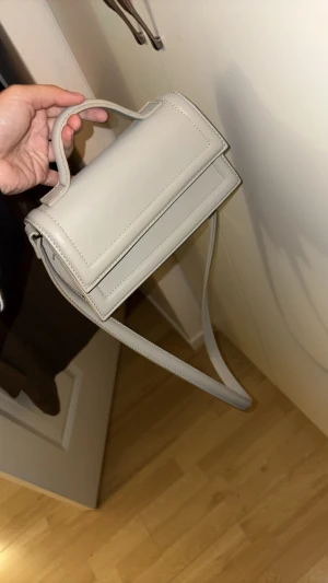 Ljusgrå axelväska med stilren design - Snygg  axelväska med minimalistisk och boxig form. Väskan har både handtag och lång axelrem, samt ett rymligt fack med dragkedja inuti. Materialet är slätt syntet och väskan har stilrena sömmar och silverfärgade detaljer. Perfekt för dig som gillar clean och modern look. Se defekt sista bild.