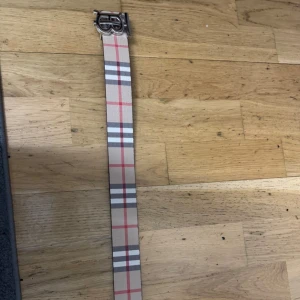 Burberry bälte  - Säljer ett snyggt Burberry-bälte med klassiskt rutigt mönster i beige, rött, svart och vitt. Bältet har en stilren silverfärgad spänne med Burberrys ikoniska TB-logga. Ena sidan är rutmönstrad, andra sidan är helsvart. Materialet är skinn.