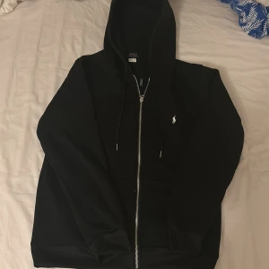 Svart hoodie från Polo Ralph Lauren - Svart hoodie från Polo Ralph Lauren med dragkedja och huva. Klassisk design med vit broderad logga på bröstet och snörning i huvan. Tillverkad i mjuk bomull, perfekt för chill dagar. Snygg och enkel att matcha med jeans eller joggers. Aldrig andvände