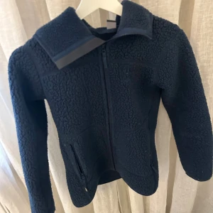 Mörkblå teddyjacka från Haglöfs - Mysig mörkblå teddyjacka med huva och hel dragkedja framtill. Jackan har två fickor med dragkedja och en skön, fluffig struktur. Perfekt för kyliga dagar och enkel att slänga på över en hoodie. Snygg och praktisk design som håller dig varm.