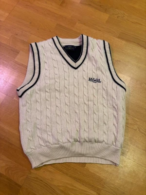 Vit kabelstickad väst från Henri Lloyd - Säljer en vit kabelstickad väst från Henri Lloyd med mörkblå detaljer vid v-ringad hals och ärmslut. Västen har broderad logga på bröstet och ribbad nederkant. Perfekt för dig som gillar klassisk och sportig stil.