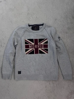 Grå stickad tröja Morris S - Snygg grå stickad tröja från Morris i storlek Small. Framsidan har en stor Union Jack-flagga med Morris-text i vinrött, svart och beige. Broderad detalj med 'MG 1968' på ena ärmen. Rund hals och ribbade muddar för en klassisk look.