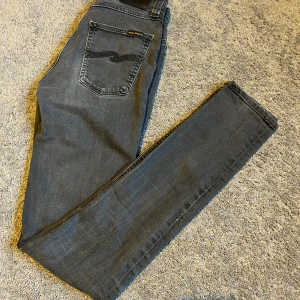 Mörkgrå jeans från Nudie Jeans, W27 L34 - Snygga mörkgrå jeans från Nudie Jeans med klassisk femficksdesign och svarta sömmar på bakfickan. Modellen är slim och raka i benen, tillverkade av ekologisk bomull med lite stretch för extra komfort. Perfekt passform och stilren look.
