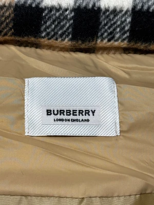 Beige pufferväst från Burberry - Snygg beige pufferväst från Burberry med ikoniskt rutigt foder i huvan. Västen har svarta dragkedjor på fickorna, tryckknappar och ett stort Burberry-märke på insidan. Perfekt för lager-på-lager och streetstyle.