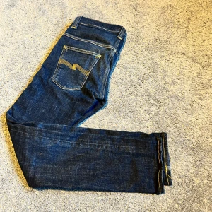 Nudie Jeans Thin Finn blå W31 L32 - Snygga blå Thin Finn jeans från Nudie Jeans med klassisk femficksdesign och kontrastsömmar. Modellen har smal passform och är tillverkad i 98% ekologisk bomull och 2% elastan för extra stretch. Perfekt för dig som gillar stilrena och bekväma jeans.