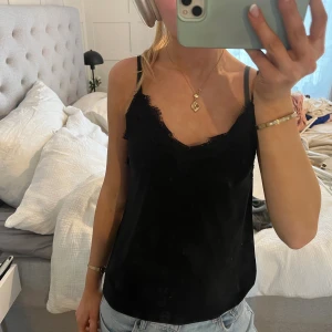 Svart linnetopp med spets från Amisu - Säljer en svart linnetopp från Amisu i storlek XXS. Toppen har tunna axelband och v-ringad hals med snygga spetsdetaljer längs kanten. Materialet är mjukt och lätt, perfekt för att styla med jeans eller kjol. Spetsen ger en extra touch av elegans.