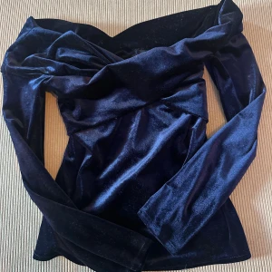Mörkblå sammetstopp från Zara Basic - Säljer en mörkblå offshoulder topp från Zara Basic Collection i mjuk sammet. Toppen har långa ärmar och en snygg omlott-detalj vid halsen som ger en elegant look. Perfekt för dig som gillar stilrena och trendiga plagg med lyxig känsla.